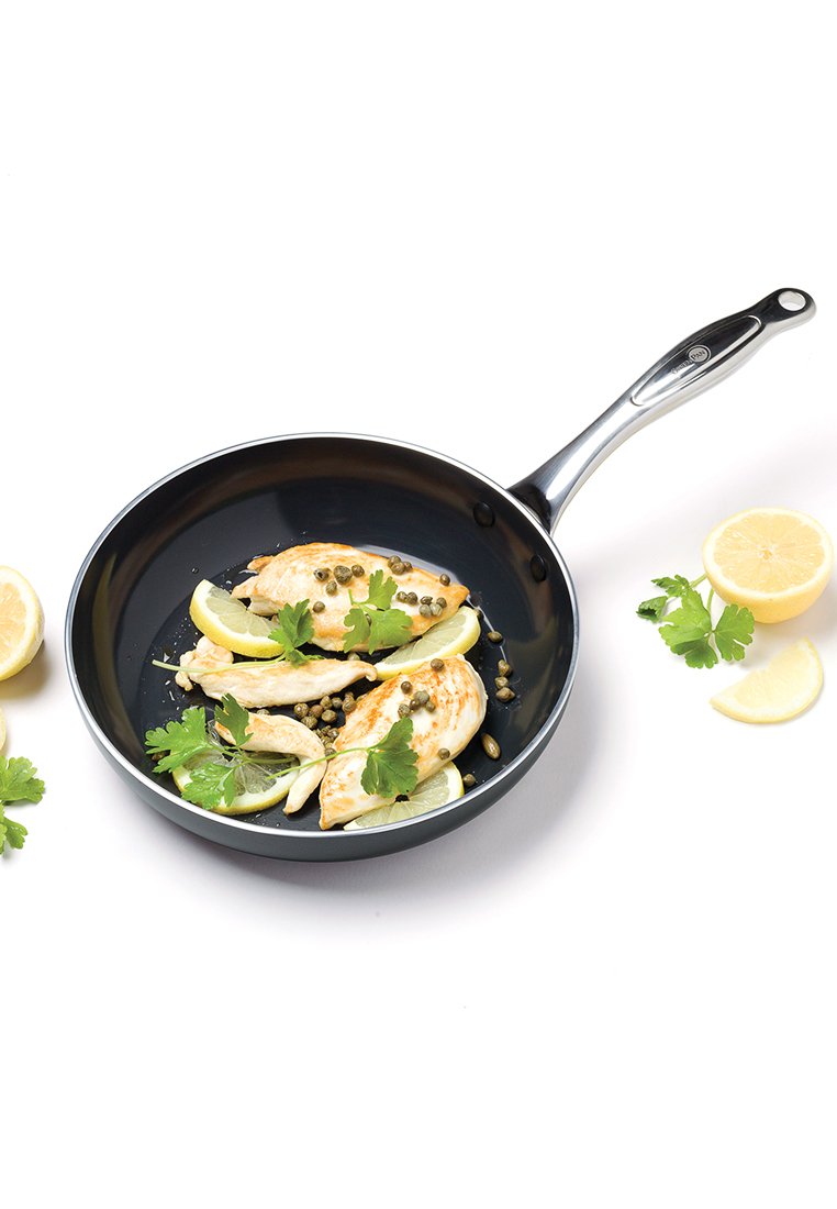 Zwarte koekenpan met gekookte kipfilets, limoenpartjes, kappertjes en peterselie op een witte ondergrond, met limoenhelften en peterselie in de buurt.