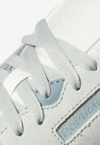 Gros plan d'une basket Reebok en daim gris clair avec des lacets plats et un logo embossé discret sur le côté près des œillets.