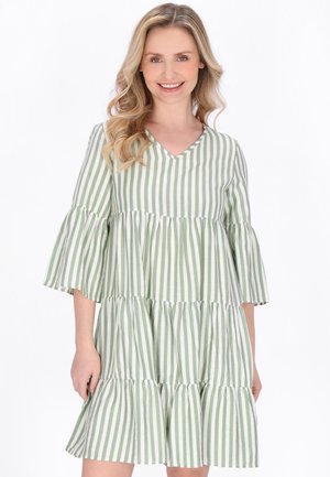 Kvinde med bølget blondt hår iført en løs, lagdelt kjole med grønne og hvide lodrette striber, smilende mod en hvid baggrund.