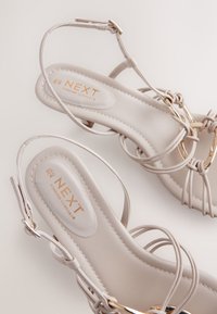 Sandales à brides beige clair avec une texture lisse, ornées d'accents dorés et de brides de cheville réglables. Marquées "NEXT Forever Comfort."