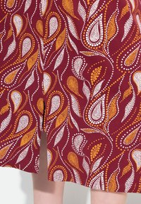 Červená tkanina se vzorem paisley v bílé a oranžové tečce, s hladkým povrchem a mírným leskem, ustřižená těsně nad kolenem.