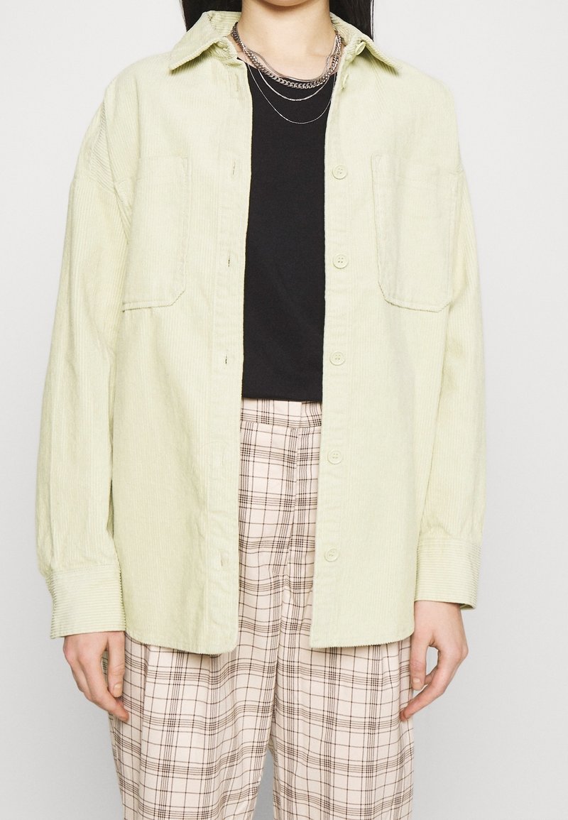 Personne portant une chemise en velours côtelé jaune clair boutonnée sur un haut noir, un pantalon beige à carreaux et des colliers en chaîne argentés superposés.