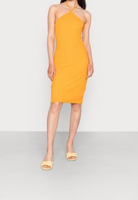 Pomarańczowa sukienka bodycon z dekoltem na halter i skrzyżowanymi ramiączkami. Materiał jest gładki i dopasowany. Uzupełniona jasnopomarańczowymi sandałami na obcasie.