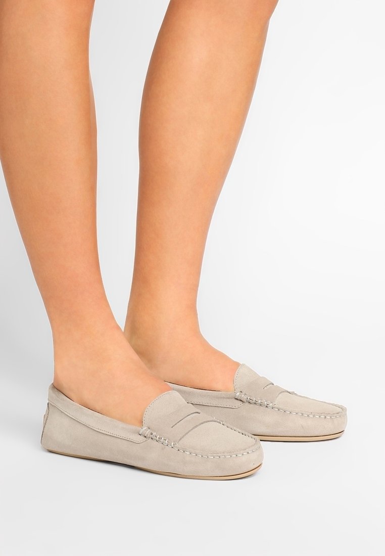 Mocassins en suede gris clair avec un bout arrondi, des coutures décoratives et sans lacets, présentés sur des jambes nues contre un fond blanc.