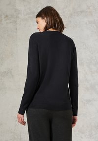 Schwarzer Pullover mit langen Ärmeln, rundem Halsausschnitt und Rippensaum. Weicher Stoff, minimalistisches Design, getragen über dunklen, schmal geschnittenen Hosen.