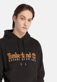 Giovane con occhi azzurri e orecchini, che indossa un felpa nera Timberland con logo arancione e bianco su sfondo bianco.