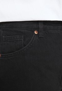 Pantalones de mezclilla negra con una textura resistente, que cuentan con un pequeño bolsillo frontal, un botón de color cobre y presillas para el cinturón estándar.