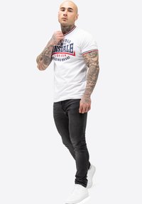 White short-sleeve polo shirt with red and blue accents, featuring a graphic logo. Paired with dark jeans and white sneakers.