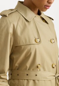 Beige trench coat, jossa on kaksoisnapitus, muoviset napit, olkaimet ja sidottu vyö; sileä kangas ja klassinen kaulus.