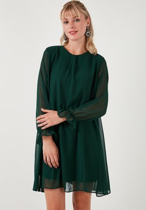 Grøn chiffonkjole med rund halsudskæring, lange gennemsigtige ærmer og flæsekantede manchetter. Let og flydende silhuet med en knælængde.