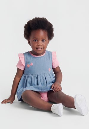 Göndör hajú kisgyermek, rózsaszín pólót és kék ruhát visel, amin cseresznyék díszelegnek, fehér cipőben ül a padlón és mosolyog.