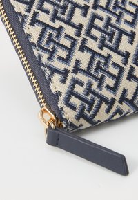 Canvas pouch in beige with navy geometric pattern, gold zipper hardware, and navy leather accent tag. Compact and structured design.