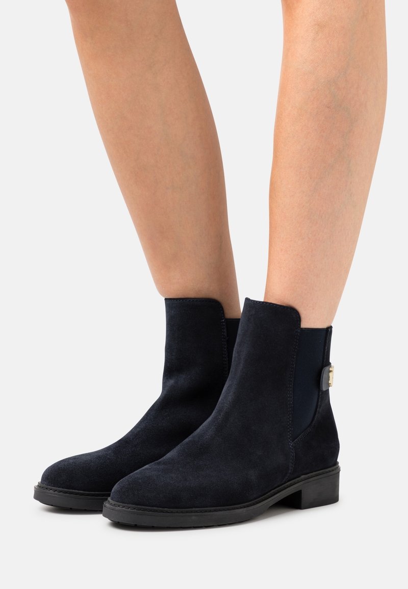 Bottes courtes en daim noir avec talons carrés bas et panneaux élastiques latéraux portées sur des jambes nues sur un fond clair.