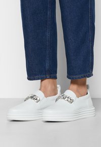 Vita slip-on skor med slät läderfinish, utrustade med en låg platåsula och en dekorativ strassdekoration framtill.