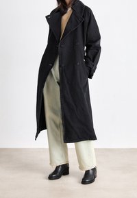 Trench coat nero doppiopetto con ampio colletto e tasche laterali, abbinato a jeans a vita alta verde chiaro e stivaletti neri.