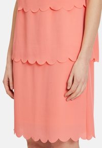 Korallenfarbenes Kleid mit geschichtetem Design, das wellenförmige, geschwungene Abschlüsse an Saum und Ärmeln aufweist. Weicher, leichter Stoff mit glatter Textur.