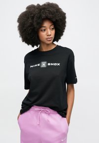 Μαύρο βαμβακερό T-shirt με το λογότυπο "NIKE SHOX" σε λευκό. Κοντά μανίκια με χαλαρή εφαρμογή, συνδυασμένο με ανοιχτό ροζ φόρμα.