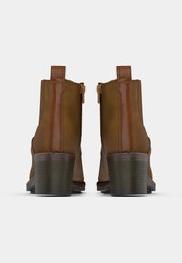 Un par de botas marrón hasta el tobillo con tacones cuadrado, vista trasera mostrando las lengüetas y cremalleras laterales contra un fondo blanco liso.