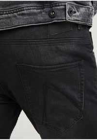 Czarne dżinsy o wyraźnym, skośnym wzorze tylnej kieszeni i kontrastowych detalach ściegów, widziane z lekko skośnego kąta.