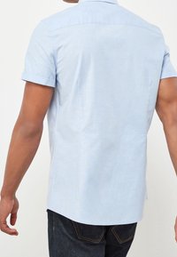 Camisa de manga corta de color azul claro, hecha de una tela suave, presenta un cuello con botones y un dobladillo curvado con costuras visibles.