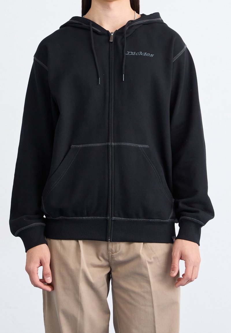 Personne portant un sweat à capuche noir zippé avec des poches avant et un pantalon beige clair, debout devant un fond uni.