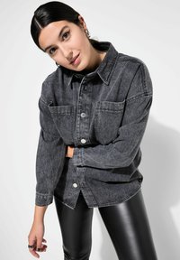 Camisa de denim gris oscuro con dos bolsillos frontales, cierre de botones y corte holgado. Combina con leggings negros y brillantes. El fondo es claro.