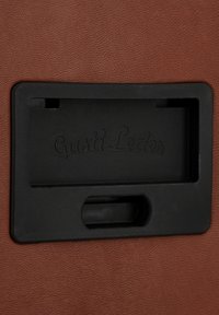 Brown textured leather background with a rectangular black plastic card holder featuring embossed text "Gusti Leder."