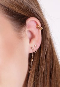 Boucles d'oreilles dorées avec des accents en forme d'étoile, un design en maillons de chaîne et de petites pierres précieuses transparentes. Plusieurs piercings visibles sur l'oreille.