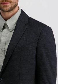 Man in een donker gestructureerde blazer over een lichtgrijze blouse tegen een plain witte achtergrond.