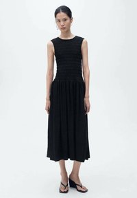 Abito nero senza maniche con corpino arricciato e gonna fluida, caratterizzato da una silhouette lunga e un tessuto testurizzato. Indossato con sandali con zeppa neri.