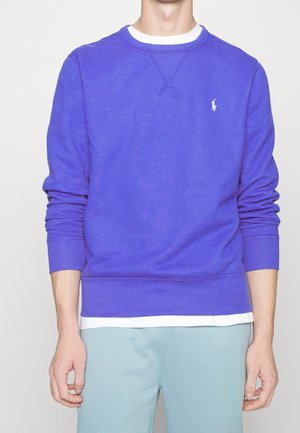 Homme portant un sweat-shirt violet à col rond avec un sous-vêtement blanc et un pantalon bleu clair, debout devant un fond uni.