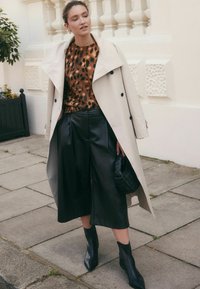 Femme portant un trench beige par-dessus un pull à imprimé léopard, un pantalon culotte en cuir noir, des bottines noires, et tenant un sac à main tissé noir en extérieur.