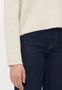 Kvinne iført en kremfarget strikkegenser og mørkeblå jeans, med venstre hånd avslappet ved siden av.