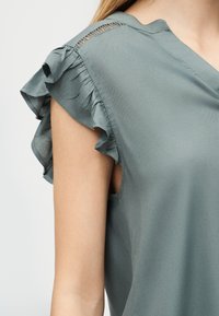 Αμάνικη γκρι μπλούζα με βολάν στο τελείωμα των ώμων και διακριτική δαντέλα κοντά στη γραμμή του λαιμού, που φοριέται από ένα άτομο με ανοιχτόχρωμο δέρμα και ξανθά μαλλιά.