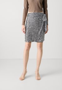 Hopeapintaiset paljettihuivi, jossa on solmittava yksityiskohta, polvipituinen, yhdistetty ruskean istuvan pitkähihasen paidan ja läpinäkyvien korkokenkien kanssa.