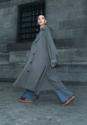 Lang grijs trenchcoat met grote zwarte knopen, gelaagd over uitlopende denimbroeken, gecombineerd met bordeauxrode schoenen met een decoratief patroon.
