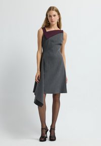 Graues figurbetontes Kleid mit einem tiefen kastanienbraunen asymmetrischen Ausschnitt. Verfügt über einen gerafften Saum und eine glatte Textur, kombiniert mit schwarzen High Heels.