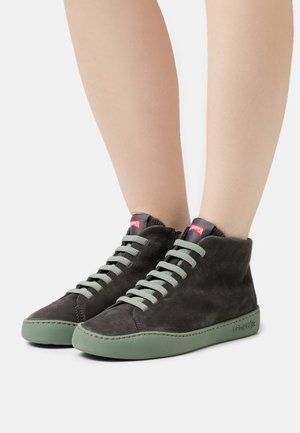 Zapatillas altas de ante negro con suelas de goma verde. Presentan cordones grises y una marca roja en la lengüeta. Textura suave, diseño minimalista.