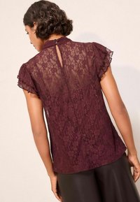 Burgundy csipke felső rövid fodros ujjal és kulcslyuk hátrésszel. Virágmintás és texturált felület jellemzi. Barnás nadrággal párosítva.