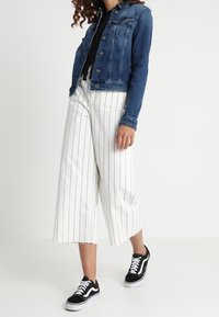 Tummansininen farkkutakki, musta pitkähiha, valkoisilla raidoilla varustetut väljät housut ja mustat tennarit, joissa on valkoiset pohjat ja kengännauhat.