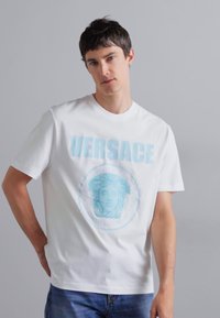 Balta kokvilnas T-kreklā ir zils "VERSACE" teksts un klasiskas figūras grafika apļveida dizainā. Īsas piedurknes, apaļa kakla izgriezums.