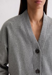 Cardigan gris à boutons avec une coupe décontractée, un col en V, une texture subtile et des coutures proéminentes le long de l'ouverture frontale.