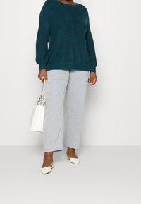 Sweter dzianinowy w kolorze morskim z długimi rękawami, jasnoszare spodnie o szerokich nogawkach oraz białe płaskie buty; trzyma białą torebkę o usztywnionej formie z metalicznymi zdobieniami.