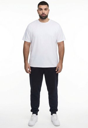 Uomo in piedi rivolto in avanti con una maglietta bianca semplice, pantaloni da jogging neri e scarpe da ginnastica bianche su uno sfondo bianco.