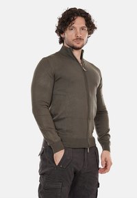 Cardigan verde oliva con zip, design aderente con texture a coste e colletto alto. Lazipper presenta un piccolo tirante in metallo. Indossato con pantaloni cargo scuri.