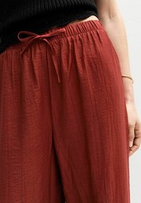 Pantalons large en couleur rouille avec un tissu texturé, dotés d'une taille élastique et d'une fermeture à cordon de serrage noué.