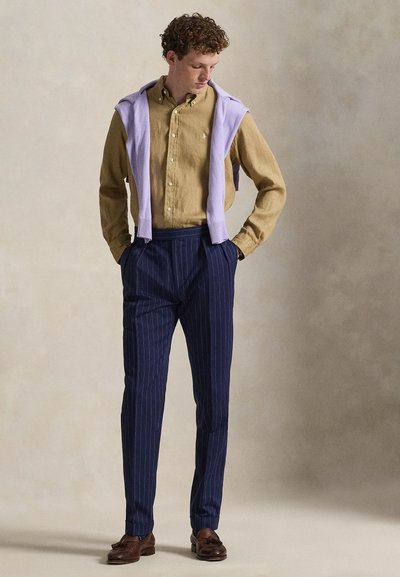 Koszula na guziki w kolorze musztardowym, w połączeniu z granatowymi spodniami w prążki. Lawendowy sweter zarzucony na ramiona, brązowe skórzane buty.