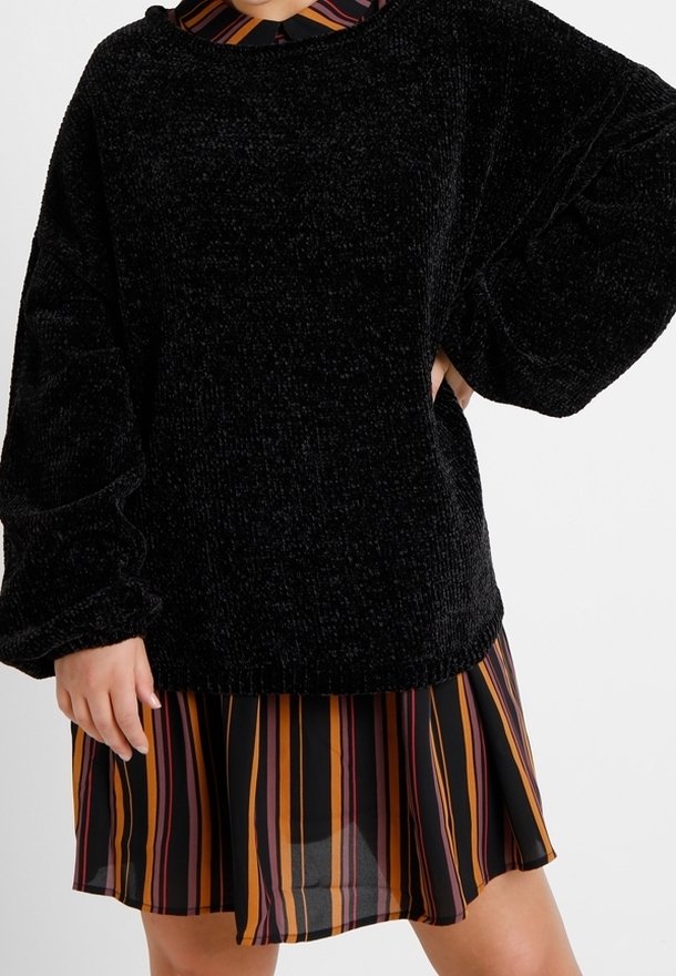 Maglione nero con texture e maniche oversize indossato sopra un vestito a strisce multicolori con linee verticali arancioni, gialle e nere.
