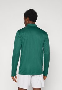 Πράσινο αθλητικό μακρυμάνικο πουκάμισο με όρθιο γιακά, λεία υφή και εφαρμοστή σχεδίαση. Φορεμένο με λευκές σορτς, από πίσω όψη.