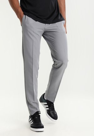 Persoon draagt strak zittende lichtgrijze pantalon en zwarte sneakers met witte strepen, staat met handen in zakken tegen een effen achtergrond.